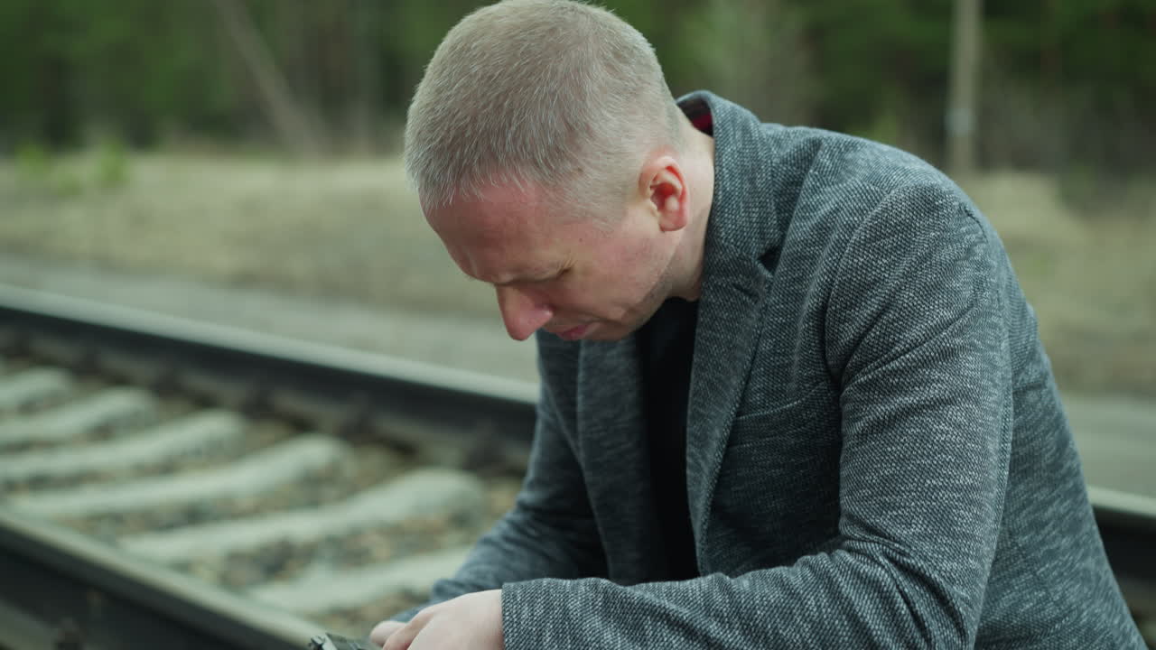 A close view of a man in a gray jacket handling a handgun while sitting down beside a railway track, intently inspecting the firearm, the background shows a blurred view of a green forest