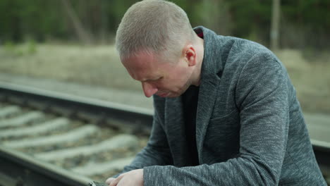 a close view of a man in a gray jacket handling a handgun while sitting down beside a railway track, intently inspecting the firearm, the background shows a blurred view of a green forest