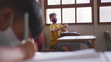 boy wearing face mask sneezing while sitting on his desk at school