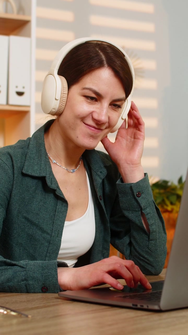 Giovane donna al tavolo di casa che lavora al laptop mentre ascolta musica con cuffie wireless all'interno