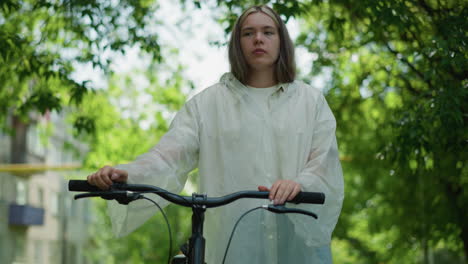 joven con impermeable translúcido se detiene mientras sostiene su bicicleta, quita la mano del manillar, y toma una respiración profunda, mirando perdido en el pensamiento, rodeado de vegetación exuberante y la suave luz del sol