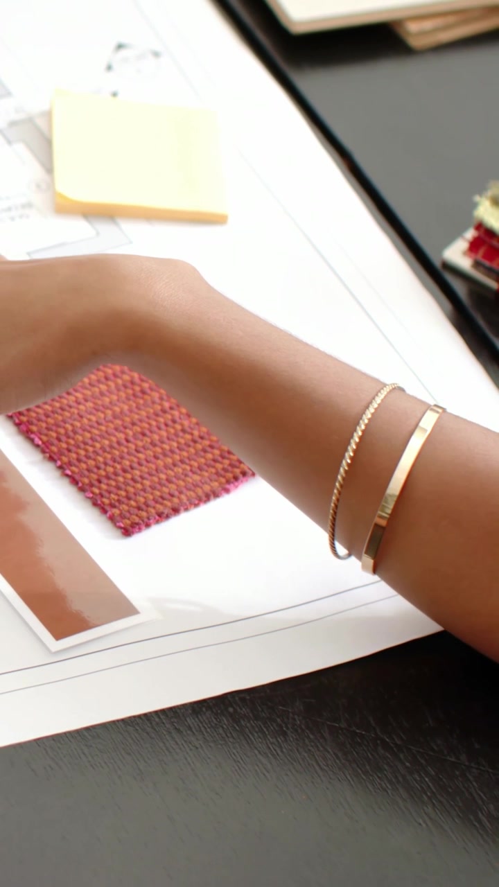 Vertical video: Two-gold-bracelet arm entering moving at desk aligning plan and red swatch choosing