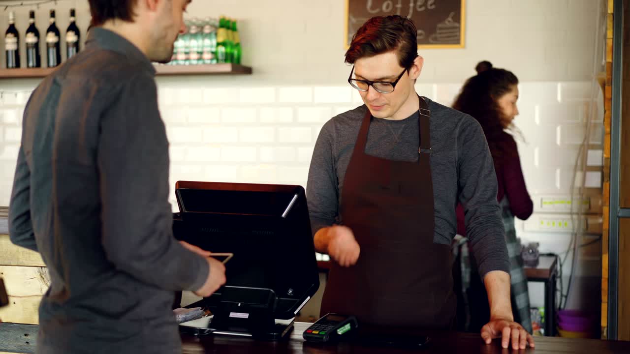 Handsome male cashier is accepting contactless smartphone payments while attractive young waitress is bringing takeaway coffee to customers in coffee-shop.