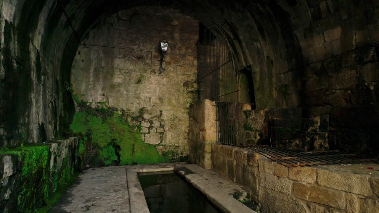 Vista interior de um túnel de pedra escuro e arqueado, com paredes cobertas de musgo e antigos bebedouros na sua base. A iluminação fraca destaca a textura desgastada da arquitetura histórica e a umidade.