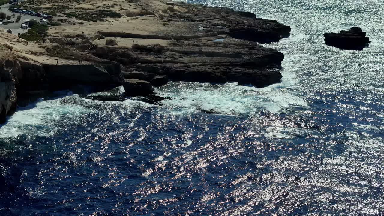 Dramatic high angle aerial view of the rugged limestone coastline of Gozo Malta during a bright sunny summer day featuring crashing deep blue Mediterranean waves against dark rocky shore cliffs