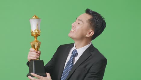 close up of asian business man in a suit and tie with a gold medal kissing a gold trophy being proud winning as the first winner on green screen background in the studio