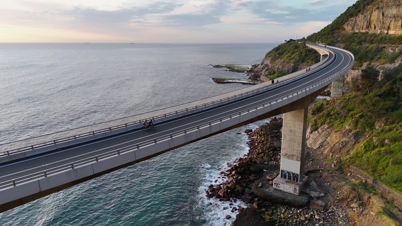 Aerial tracking shot of a bicycles along the iconic Sea Cliff Bridge on the Grand Pacific Drive, Australia, with stunning drone coastal views