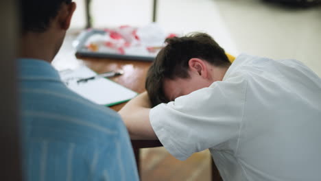 back angle of white lad resting head on folded arm while leaning on wooden desk ambient indoor light capturing peaceful posture with tousled hair slight blur creating intimate calm mood soft glow beam