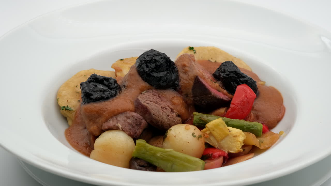 Food on a plate Meat dumplings potatoes and green beans are mixed with brown sauce and topped with dark fruit in a white bowl