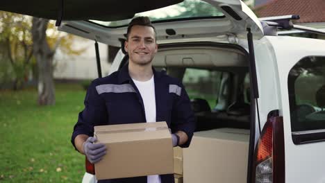 hombre sonriente guapo en uniforme sacando la caja del maletero y mirando a la cámara. entrega de paquetes, carrito de mano