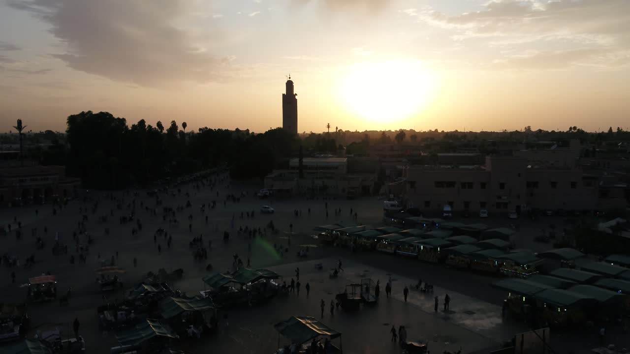 Fas, Marakeş'teki Camiü'l-Fena Meydanı ve Kutubiye Camii siluetinin sinematik gün batımı manzarası, canlı turuncu bir gökyüzü ve şehir kalabalığı eşliğinde