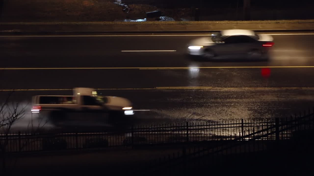 Cars glide along a rain-soaked road at night, street lamps reflecting on the wet pavement and casting a moody, cinematic glow over the scene