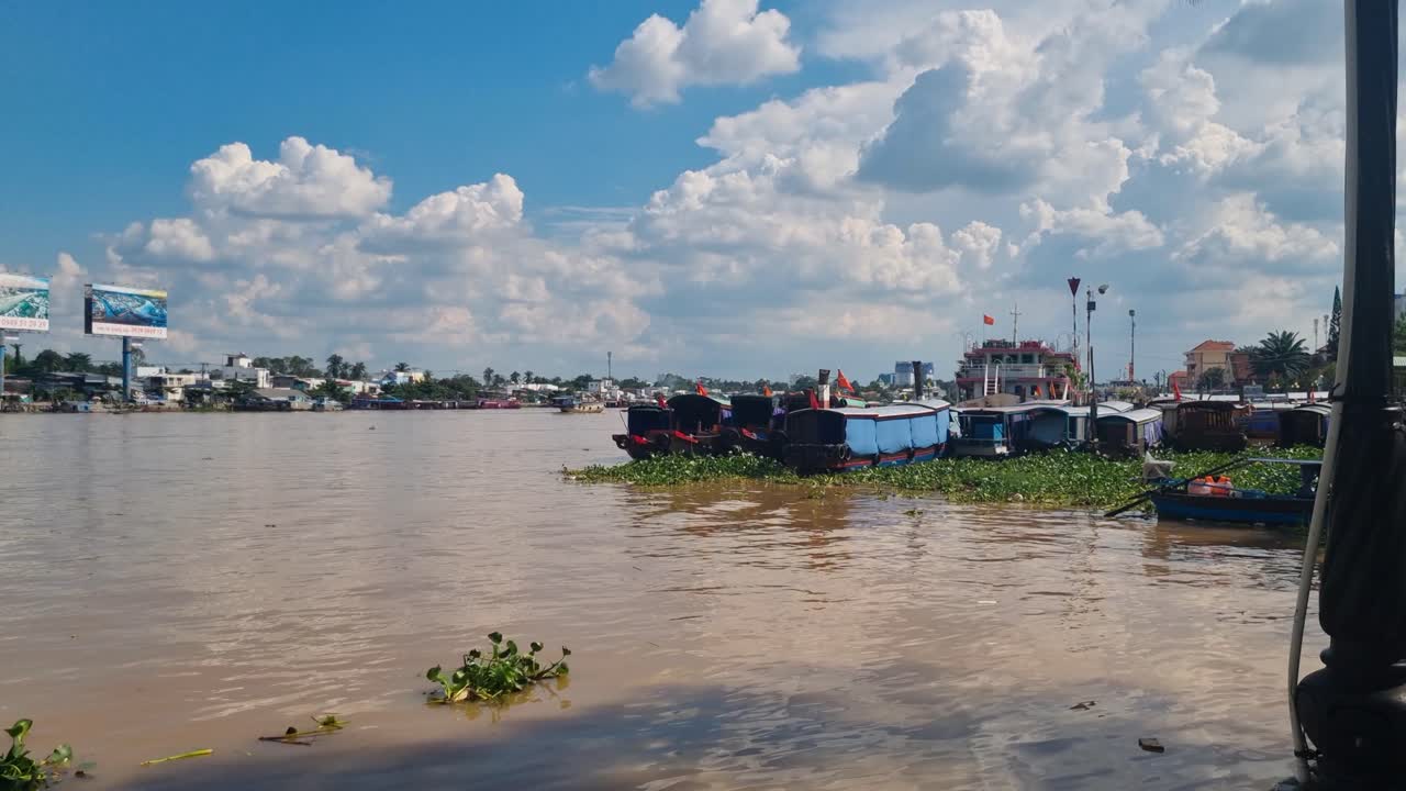 River view in Can Tho, Vietnam: wooden boats are anchored on the Hau (Bassac) River, floating plants drift beneath a blue sky with white clouds, reflecting calm daily life in the Mekong Delta