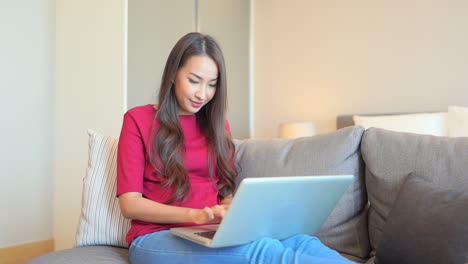 Mujer-Asiática-Sentada-En-El-Sofá-Con-Su-Laptop-En-El-Regazo-Y-Escribiendo-En-El-Teclado-Mostrando-La-Hermosa-Sonrisa