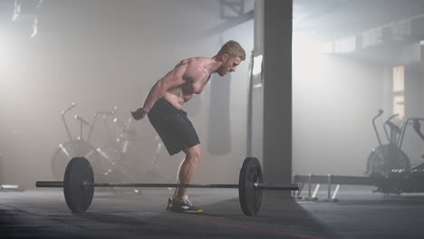 a young man without a shirt in slow motion jumps a burpee over a barbell in the gym in backlight