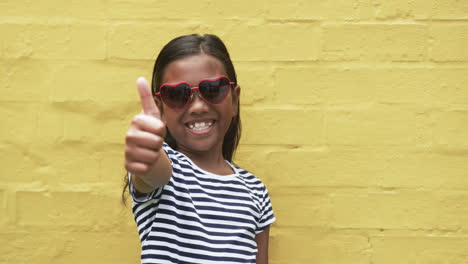 a young biracial girl stands before a yellow background, giving a thumbs up