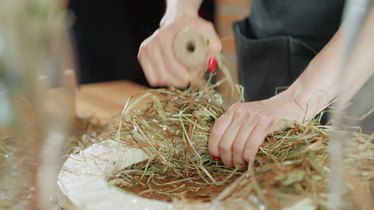 Decorator arranging hay with spool of twine while crafting rustic wreath on wooden table, showing closeup of skilled hands with red nails working carefully to create handmade seasonal decoration