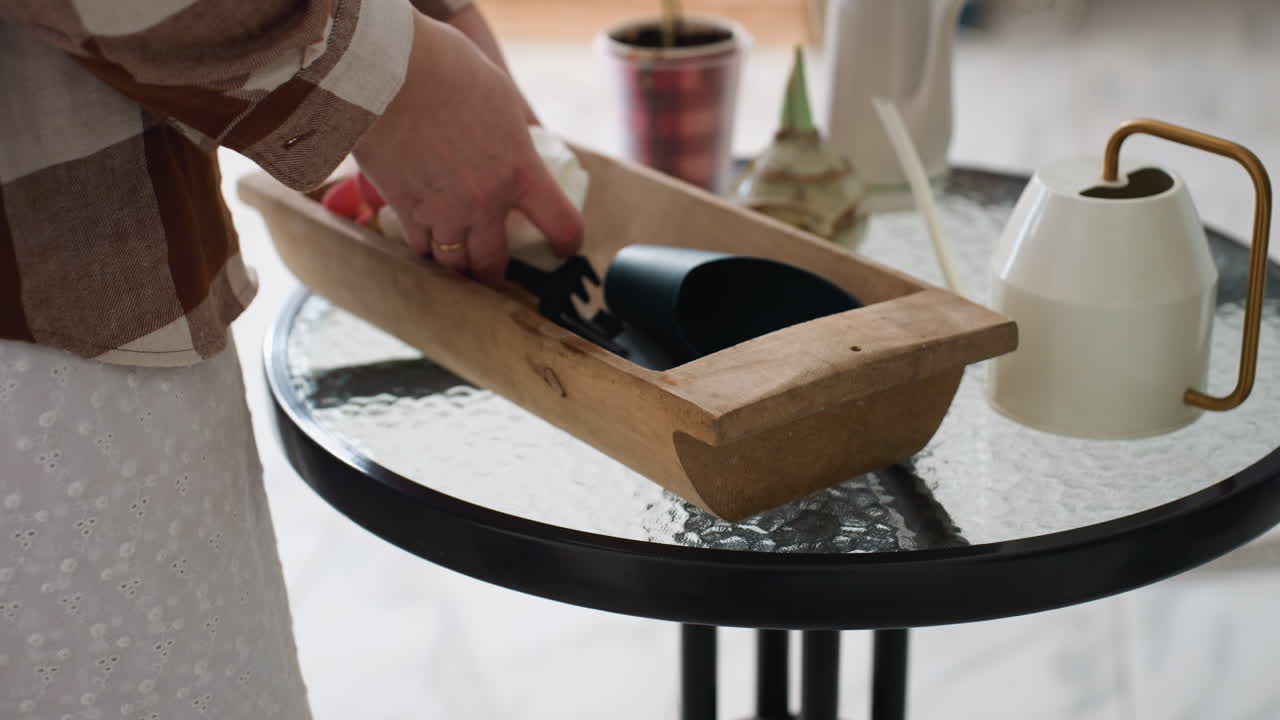 Farmer hands arranging tools in wooden container on round glass table near potted plants in winter home interior with soft natural light streaming through sheer curtains creating inviting scene
