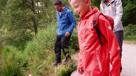 Familia-Joven-Caminando-Hacia-Un-Lago-Y-Jugando-Juntos-En-Una-Rama-Caída,-Lake-District,-Reino-Unido