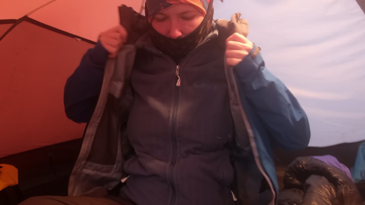 A girl in a tent sits and puts on a membrane jacket before climbing