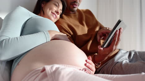 expecting couple relaxing on bed, using tablet and enjoying time together