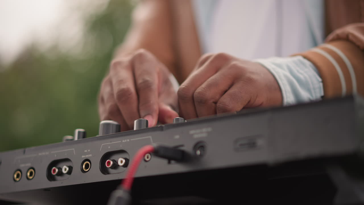 Nærbillede af sorte DJ-hænder, der justerer mixer-kontroller, fingre drejer knapper og tilslutter et rødt kabel, opsætning i udendørs park med varmt aftenlys, fokuseret sjælfuld stemning, taktil fingerspidsbevægelse, bærbar