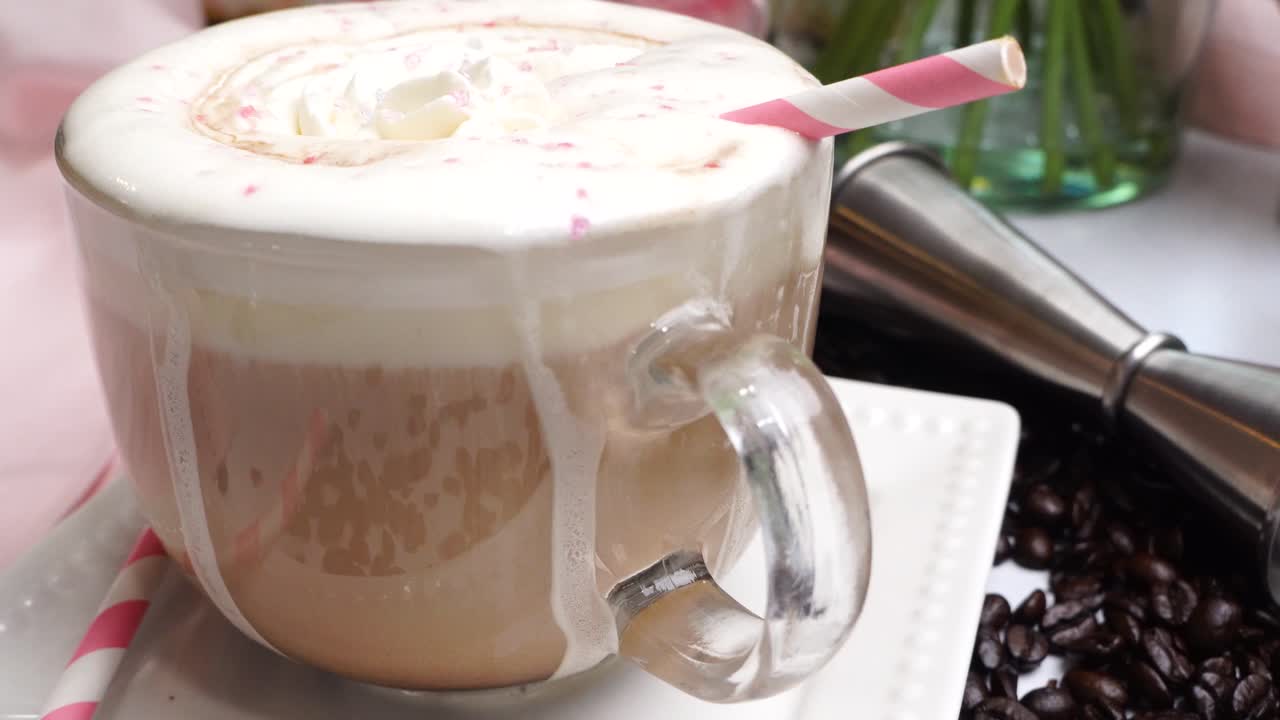 Dessert pink coffee in a mug topped with whipped cream and sugar sprinkles. Delicious and sweet dessert hot coffee in a coffee mug with a pink striped straw and surrounded by coffee beans.