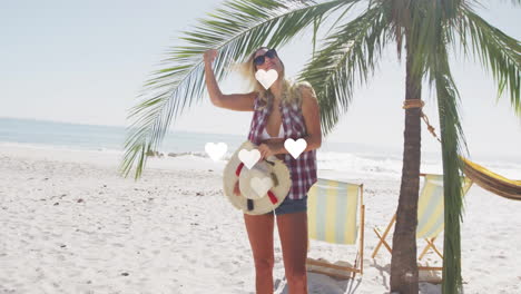 corazones animación sobre mujer sosteniendo sombrero y de pie cerca de la palmera en la playa