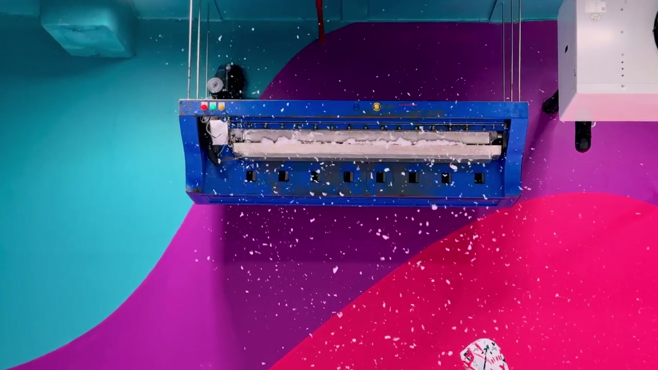 Una macchina per la neve artificiale blu sospesa al soffitto rilascia fiocchi bianchi all'interno di un centro divertimenti al coperto dai colori vivaci. Il dispositivo meccanico crea una divertente atmosfera invernale su pareti vivaci.