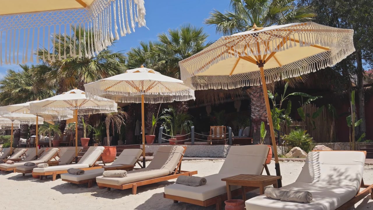Inquadratura statica grandangolare di lettini di lusso e ombrelloni boho chic con frange allineati sulla sabbia bianca in un esclusivo resort sulla spiaggia tropicale sotto un cielo azzurro e limpido.