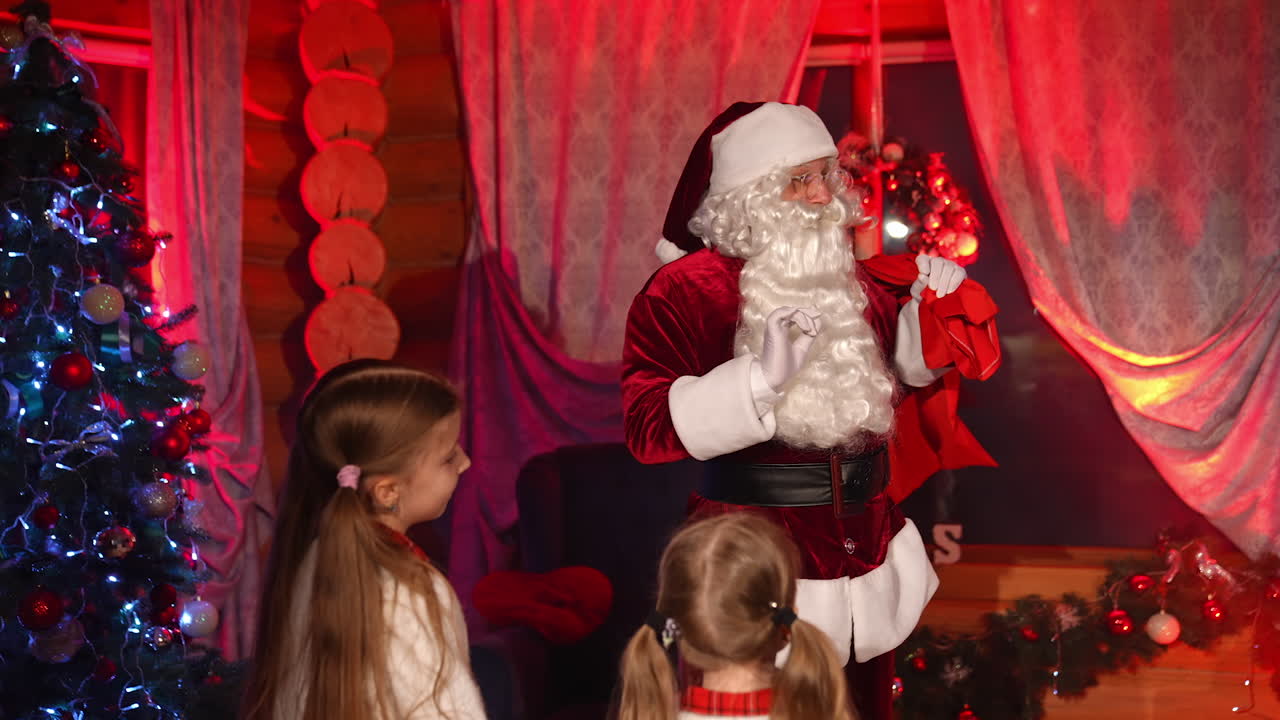 Santa Claus with children. Santa Claus with white beard and red suit in a house holding sack with Christmas gifts and talking to little girls. Family celebration. Red lighting.