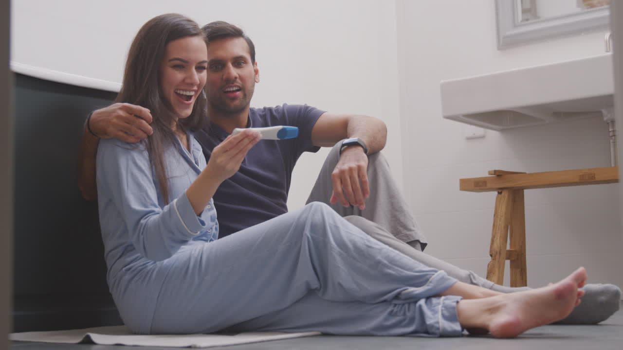 Pareja emocionada sentada en el suelo del baño de casa con una prueba de embarazo casera positiva