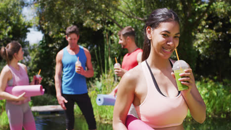 mujer biracial sonriente bebiendo bebida de salud, con grupo diverso hablando después de yoga en el parque soleado