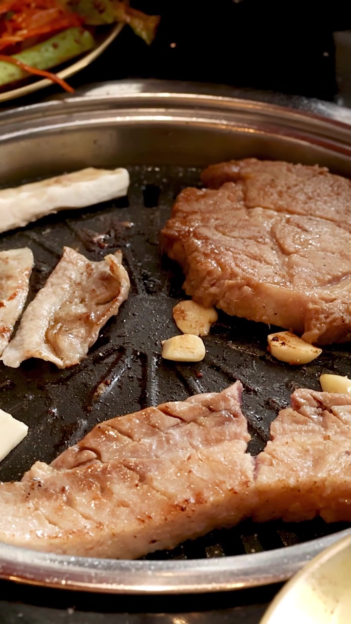 Irisan daging babi dan bawang putih dipanggang di panggangan meja, pencahayaan hangat, jarak dekat, tangan menggunakan penjepit.