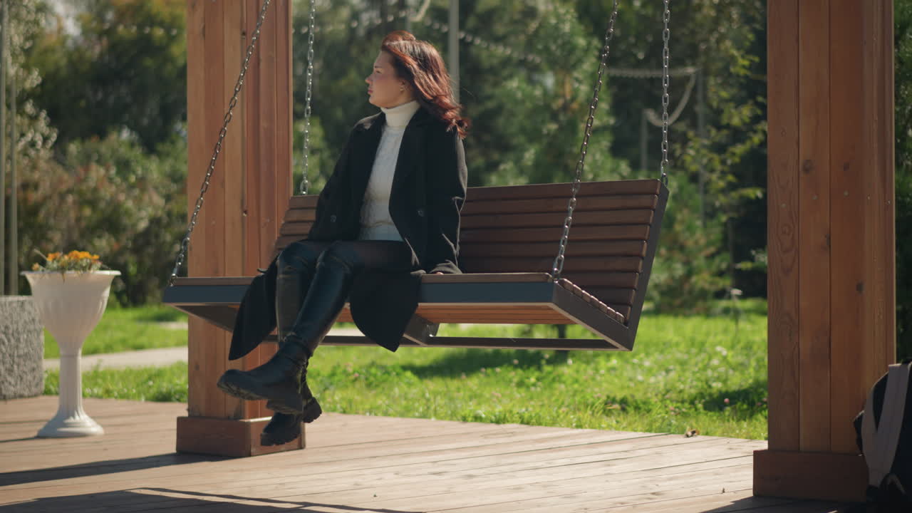 검은 코트와 가죽 부츠를 입은 여인이 공원에서 나무로 된 스 의자에 앉아 있습니다. 머리카락은 바람에 흔들리고, 가방은 나무 기둥에 누워 있습니다. 울창한 초록색으로 둘러싸여 있습니다.