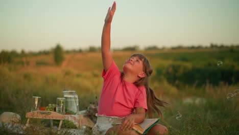 young girl in pink top seated outdoors in nature, joyfully reaching up to catch floating bubbles with a playful expression, surrounded by greenery, picnic setup