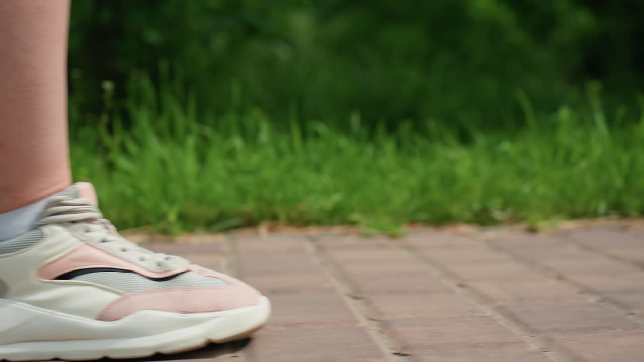 녹색 공원에서 햇빛을 받으며 벽돌길을 따라 걷는 여성의 옆모습 다리 부분을 클로즈업하여 촬영한 사진이에요. 자연과 풀로 둘러싸인 포장 도로에서 운동화의 움직임이 잘 드러나요.
