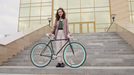 Smiling-Curly-Woman-In-Formal-Clothes-Looking-At-The-Camera-While-Standing-On-The-Stairs-With-Her-Bike