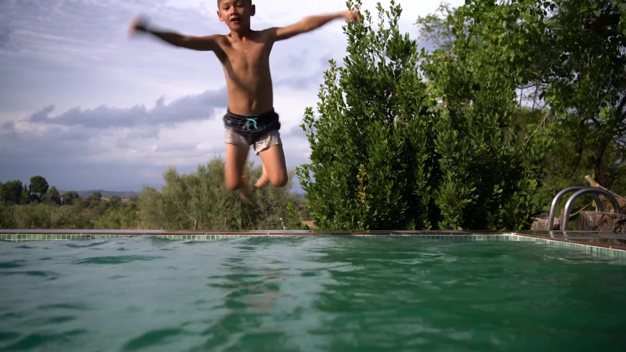 Un ragazzo sta in piedi sul bordo di una piscina a sfioro con le braccia distese prima di tuffarsi e scomparire sotto uno schizzo, Inquadratura statica.