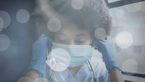 Animation-of-glowing-light-spots-over-smiling-african-american-female-doctor-wearing-face-mask