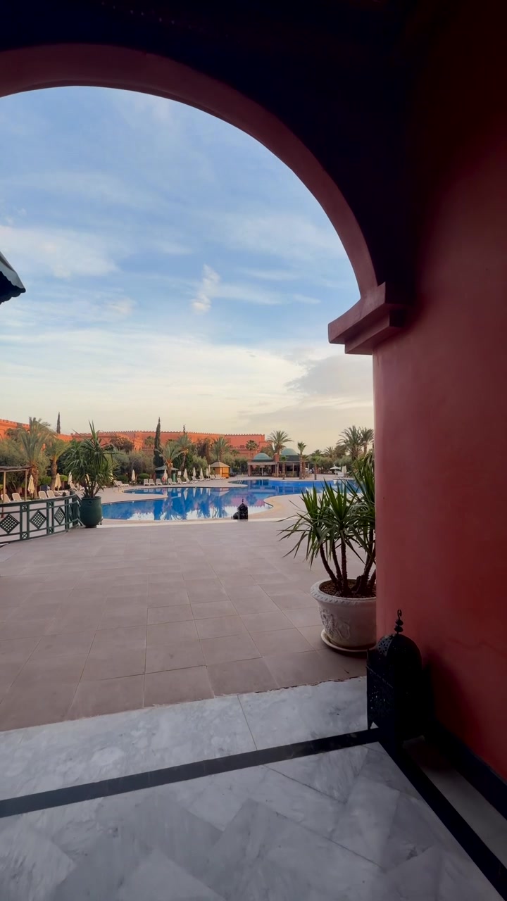 Vista attraverso un arco verso la piscina di un resort con palme e paesaggio desertico, Marocco.