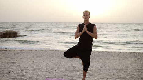 a young man is doing yoga on a sports mat, standing on one leg during the dawn of the sun. doing a specific exercise. meditation, hands. leisure time, recreation