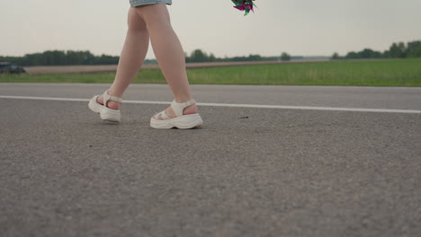 leg view of white woman walking on tarred road holding colourful pinwheel with blur view of parked car in distance beside green verge under soft summer light conveying calm motion