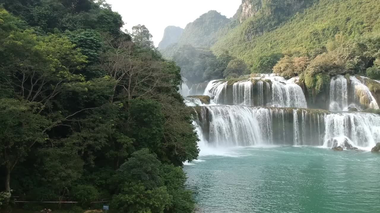 Aerial Behind Tree Reveal Of Cascading Waterfalls Of Ban Gioc With Rainbow In Vietnam. Follow Shot