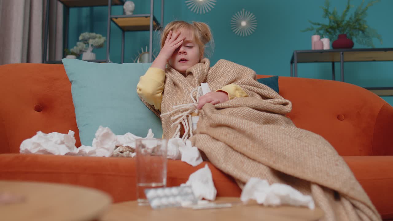 Child girl in plaid suffering from coronavirus or allergy sneezes snot into napkin on sofa at home