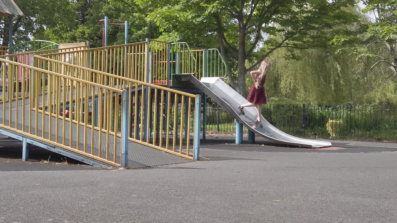 イギリス、ペイントンにあるビクトリア公園の金属製すべり台を子供が駆け下りる様子を、自然光と滑らかな動きで捉えた静止スローモーションショット。遊びの楽しさと活気を表現しています。