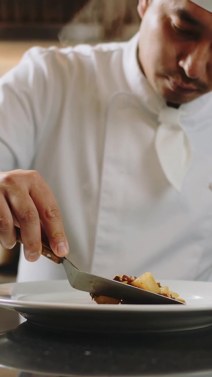 Vertical video: Plating chef in coat adjusting diced food on pass plate, steam urging spatula work