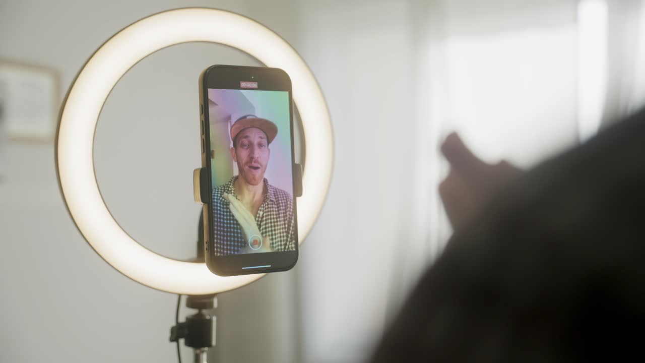 Indholdsskaber optager video. Influencer filmer med smartphone i ringlys, iført baseballkasket og ternet skjorte. Indhold til engagement på sociale medier og fans. Digital forretning streamer online.