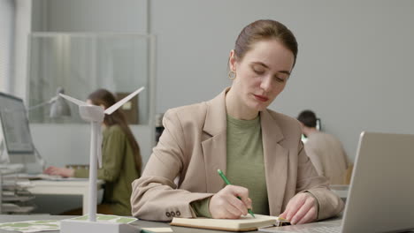 mujer usando laptop y escribiendo notas sentada en la mesa con modelo de molino de viento en la oficina