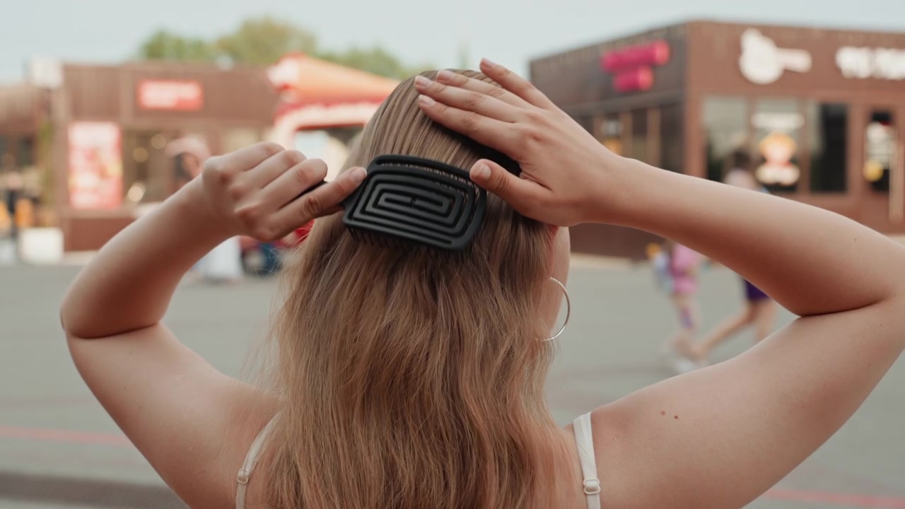 Donna con i capelli lunghi si acconcia le onde sciolte in uno spazio pubblico all'aperto, usando un pettine nero per lisciare e sistemare le ciocche. Lo sfondo presenta vetrine sfocate, pedoni, abbigliamento casual, caldo.